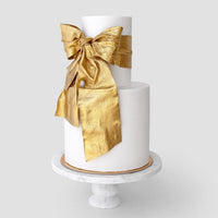 A two-tiered white cake on a marble stand, decorated with a shimmering antique gold bow made using Jenna Rae Cakes Antique Gold Edible Lustre Dust - 5g Shaker (Package of 6).
