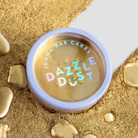 A round container from the Jenna Rae Cakes Dazzle Dust Best Seller Trio rests on a surface dusted with gold powder and liquid splatters, featuring a lavender rim and holographic text on its lid.