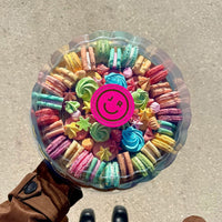 A person in a brown coat and black boots holds a Jenna Rae Cakes Assorted Party Platter—round box of colorful macarons and meringues with a clear lid featuring a pink sticker with a smiling face logo, viewed from above.