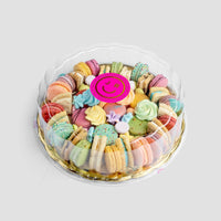 The Jenna Rae Cakes Assorted Party Platter features colorful macarons and meringue cookies in a clear round container, topped with a pink smiley-face sticker.