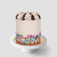 Celebrate with the Birthday Oreo Cakes 101 In-Person Cake Decorating Class by Shop Jenna Rae Cakes—learn to craft a tall cake with speckled frosting, creamy dollops, chocolate cookies, sprinkle decor, all presented on a white marble stand.