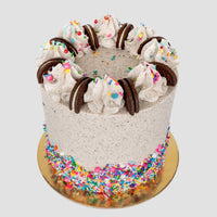 Shop Jenna Rae Cakes Birthday Oreo Cakes 101 In-Person Cake Decorating Class features a tall cookies and cream cake with swirled frosting, chocolate sandwich cookies, colorful sprinkles, and a sprinkle-covered base on a gold board.