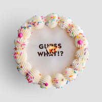 The Burn Away Cake by Jenna Rae Cakes is a round cake with white frosting, colorful sprinkles, pearl decorations, and GUESS WHAT!? written on top with a small orange heart partially covering the middle word.