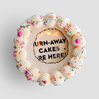 A round Burn Away Cake by Jenna Rae Cakes with white icing, colorful sprinkles, and pink and silver pearls. The center features a cutout revealing the hidden message: TURN-AWAY CAKES ARE HERE!.