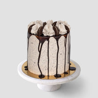 The Chocolate Oreo Cake by Jenna Rae Cakes is a tall cake topped with swirls of cream, chocolate cookies, and chocolate ganache drips, displayed on a white marble cake stand against a plain gray background.