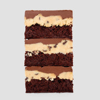 Cookie Dough Brownie by Jenna Rae Cakes features three stacked chocolate brownies topped with chocolate chip cookie dough and a chocolate glaze, displayed on a plain light gray background.