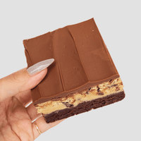 A hand with glittery nail polish holds a layered dessert bar featuring a brownie base, a cookie dough center with chocolate chips, and a smooth chocolate topping.
