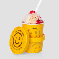 A yellow Jenna Rae Cakes Cookie Dough Cup labeled Limited Edition shows a smiley face and is filled with cookie dough, red syrup, white icing, a cherry, and a white spoon. A second identical cup and its lid rest nearby on a gray background.