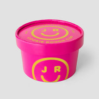 A bright pink Jenna Rae Cakes Cookie Dough Cup with a matching lid displays yellow smiley faces and JR lettering. The lid label says COOKIE DOUGH CUP in yellow. Packaging is shown on a plain, light gray background.
