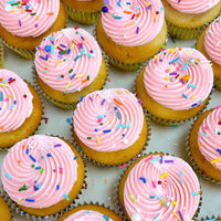 Rows of vanilla cupcakes with pink swirled frosting and colorful sprinkles, arranged on a white surface, by Jenna Rae Cakes for their Dessert Tables.