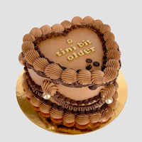 The Espresso Martini Heart Cake by Jenna Rae Cakes features piped chocolate frosting, gold pearls, and a tini bit older written in gold atop a heart-shaped cake on a gold cake board, set against a light background.