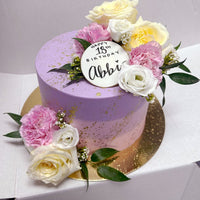 The Fresh Floral Buttercream Cake by Jenna Rae Cakes features a round purple design with gold splatters, pink and white flowers, green leaves, and a white disc on top reading Happy 18th Birthday Abbi in black. Presented on a gold board.