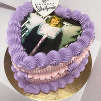 A heart-shaped cake with purple and pink frosting features a printed photo of a child in a fairy costume. A white heart topper on top reads Happy 40th Birthday Stephanie. The cake sits on a gold base.