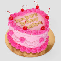 The Heart Cake by Jenna Rae Cakes is a pink, heart-shaped cake with piped pink icing, six cherries on top, and gold happy bday babe letters. It sits on a gold board against a plain light background.