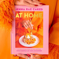 A person in an orange outfit with feathered sleeves holds the Jenna Rae Cakes at Home Cookbook by Jenna Rae Cakes, which features a dessert and gold text on the pink cover. Their nails are painted red and they wear multiple rings.