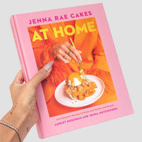 A hand with light nail polish holds the Jenna Rae Cakes at Home Cookbook by Jenna Rae Cakes, featuring a dessert on a plate and a person in an orange outfit using a gold fork on the cover.