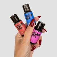 A hand with long red nails holds three Jenna Rae Cakes x Colour Mill Pack bottles from Jenna Rae Cakes in rust, blue, and pink shades against a plain light gray background.