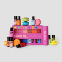 The Jenna Rae Cakes x Colour Mill Pack by Jenna Rae Cakes features multiple small food coloring bottles arranged in and around a pink box, displayed with colorful macarons and fondant shapes on a light gray background.