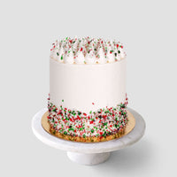 The Candy Cane Cake by Jenna Rae Cakes is a tall, round cake with smooth white frosting and red and green sprinkles on the bottom and top, shown on a white marble cake stand against a light gray background.