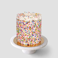 The Last Minute Cakes by Jenna Rae Cakes features a round cake covered in rainbow sprinkles and swirls of white frosting, set on a gold cake board atop a white marble stand against a light gray background.