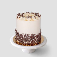 A tall, round cake with smooth white frosting, topped with piped swirls and chocolate sprinkles, and decorated with chocolate shavings around the sides. The cake sits on a white marble stand against a plain background.