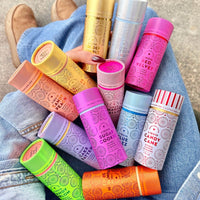 A person in blue jeans holds 12 colorful cylindrical containers of Jenna Rae Cakes Liquid Candy Peach, each with patterned designs and dessert-themed labels; Red Velvet, Candy Cane, Butterscotch, and Salted Caramel are among the flavors. Brown shoes are partly visible.