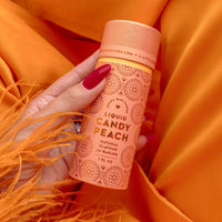 A hand with red nail polish holds a peach-colored Jenna Rae Cakes Liquid Candy Peach bottle, featuring a floral design, against vibrant orange fabric. The product is specially made for baking.