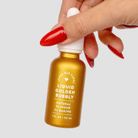 A hand with long red nails holds a small gold bottle of Jenna Rae Cakes Liquid Golden Bubbly against a plain light background.