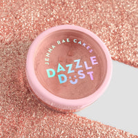 A package of six Jenna Rae Cakes Lucky Penny Edible Glitter 5g shakers, with pink lids and holographic labels, sits on a sparkling pink glitter surface reflecting various colors.