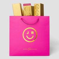 A pink Jenna Rae Cakes shopping bag with a gold smiley face, holding Macaron Gift Boxes, each decorated with matching gold smiley faces and JR letters.