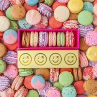 An open yellow Jenna Rae Cakes Macaron Gift Box with a pink interior displays six colorful, sprinkle- and icing-decorated macarons. The box features smiley faces and JR branding, surrounded by assorted macarons.