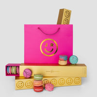 A bright pink Jenna Rae Cakes gift bag with a gold smiley face logo sits among yellow Macaron Gift Boxes filled with colorful macarons, some stacked and some scattered, all set against a white background.