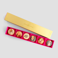 The Macaron Message Gift Box by Jenna Rae Cakes is a rectangular gold box with six round macarons, each topped with a custom photo or message like Happy Birthday Leon! The bright pink interior adds a festive touch.