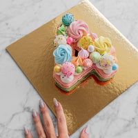 A manicured hand reaches for a Macaron Number Cake by Jenna Rae Cakes, shaped like a colorful letter A and decorated with pastel meringues and sprinkles on a gold board atop marble.