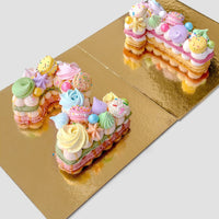The Jenna Rae Cakes Macaron Number Cake features cakes shaped into the number 21, topped with colorful meringues, macarons, candies, and sprinkles, and elegantly displayed on a gold board.