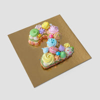 The Jenna Rae Cakes Macaron Number Cake, shaped as the number 2 and topped with pastel meringues, sprinkles, and candies, is presented on a square gold board against a light gray background.