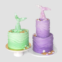 Mermaid Cakes by Jenna Rae Cakes: one green, single-tiered and one tall, purple, two-tiered—both topped with mermaid tails and decorated with wave-like frosting, seashells, and edible pearls.