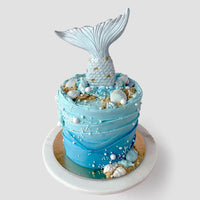 The Mermaid Cake by Jenna Rae Cakes is a round blue cake adorned with edible seashells, pearls, sand-like crumbs, and topped with a large silver mermaid tail. Presented on a white and gold cake board.