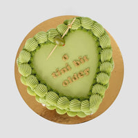 The Olive Martini Heart Cake by Jenna Rae Cakes is a heart-shaped green cake with piped frosting edges, topped with a green olive on a toothpick and a tini bit older written in gold letters, set on a gold cake board.