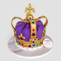 A One-of-a-Kind Cake by Jenna Rae Cakes shaped like a royal crown, adorned with gold accents and edible jewels on purple fondant, sits on a white board with a “Congrats MEC, Senior Crown Attorney” tag.