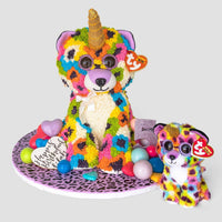 A vibrant unicorn bear cake from Jenna Rae Cakes’ One-of-a-Kind Cakes, featuring a gold horn, rainbow spots, and sunglasses, sits beside a matching Ty-tagged plush. The display includes balloons and a Happy Birthday message.