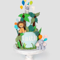 A two-tiered jungle-themed cake with fondant animals—a lion, zebra, and elephant—colorful balloons, green leaves, and the name laith on the side. The cake sits on a white stand with a number 1 topper.