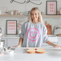 A smiling woman stands in a modern kitchen behind four slices of cake, wearing a gray sweatshirt with a pink smiley face and JR. The background features cake-themed decor and an OH LA LA sign, highlighting Jenna Rae Cakes Online Classes.