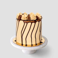 The Peanut Butter Cup Cake by Jenna Rae Cakes is a tall, round cake with light brown frosting, chocolate drip, and swirls of frosting and chocolate pieces on top, displayed on a white marble cake stand against a gray background.