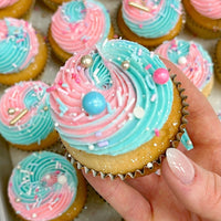 A hand holding a vanilla cupcake with swirled pink and blue frosting, topped with colorful sprinkles and edible pearls. More similarly decorated cupcakes are visible in the background.
