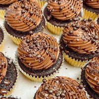 Chocolate cupcakes in yellow wrappers topped with swirls of chocolate frosting, chocolate sprinkles, and small chocolate pearls, arranged closely together on a white surface.