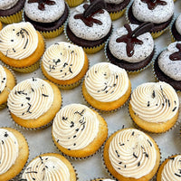 Rows of cupcakes on a tray; the front rows have yellow cake with swirls of creamy frosting topped with lavender, and the back rows have chocolate cupcakes with white frosting and a drizzle of chocolate.