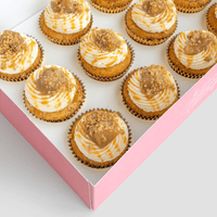 A pink box of twelve Regular Cupcakes by Jenna Rae Cakes, each topped with swirled white and caramel frosting and crumb topping, arranged neatly in rows.