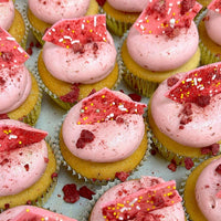 Regular Cupcakes by Jenna Rae Cakes are arranged in neat rows on a light background, each with a light yellow base, pink frosting, red candy shards, sprinkles, and crumbled red toppings.