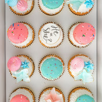 The Reveal Works Cupcakes by Jenna Rae Cakes feature 12 cupcakes topped with pink and blue macarons, pastel flowers, sprinkles, and a center cupcake reading “He or She?” for a charming gender reveal.